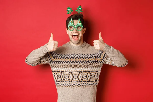 Изображение взволнованного красивого парня в очках и рождественском свитере, показывающего большие пальцы вверх, восхваляющего великий Новый год, стоящего на красном фоне — стоковое фото