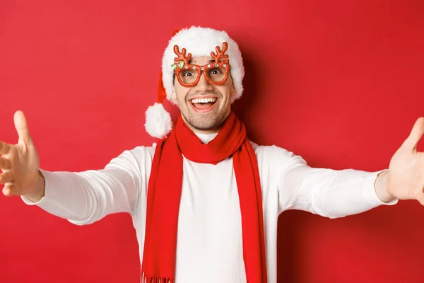 Conceito de Natal, férias de inverno e celebração. Close-up de feliz homem sorridente em óculos de festa e chapéu de Papai Noel, mãos estendidas para os lados para abraços, cumprimentando os convidados, fundo vermelho — Fotografia de Stock