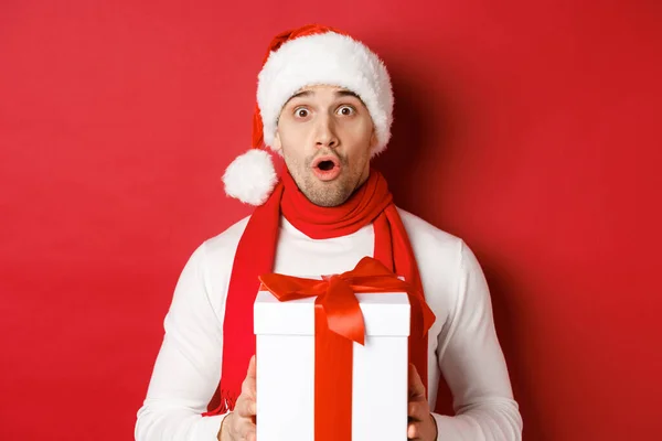 Konzept für Winterurlaub, Weihnachten und Lifestyle. Nahaufnahme eines überraschten, gutaussehenden Mannes mit Weihnachtsmütze und Schal, der erstaunt schaut und ein Neujahrsgeschenk in der Hand hält, vor rotem Hintergrund stehend — Stockfoto