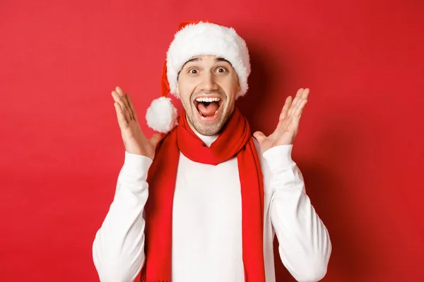 Concepto de Navidad, vacaciones de invierno y celebración. Imagen de hombre guapo mirando sorprendido en la oferta de promoción de año nuevo, sonriendo sorprendido, de pie sobre fondo rojo —  Fotos de Stock
