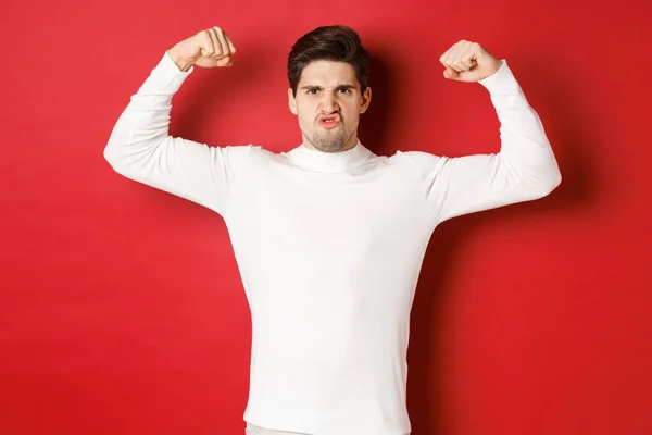 Portrait of handsome and funny guy in white sweater, flex biceps and looking encouraged, showing strong muscles, standing over red background — Stock Photo, Image