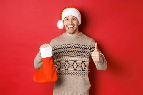 寒假的概念，新年和庆祝活动。头戴桑塔帽，头戴毛衣，英俊而快乐的男人，穿着印有糖果和礼物的圣诞长袜，竖起大拇指 — 图库照片