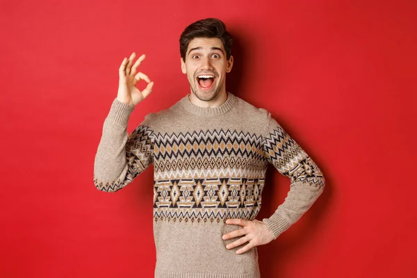 Begrepet julefeiring, vinterferier og livsstil. En ustyrlig og lykkelig mann i strikkegenser som viser tegn og ser på noe fantastisk, rød bakgrunn. – stockfoto
