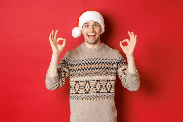 Portret szczęśliwego i zadowolonego przystojniaka, ubrany w zimowy sweter i czapkę Mikołaja, z dobrymi znakami, chwała dobrej imprezie świątecznej, stojący nad czerwonym tłem — Zdjęcie stockowe