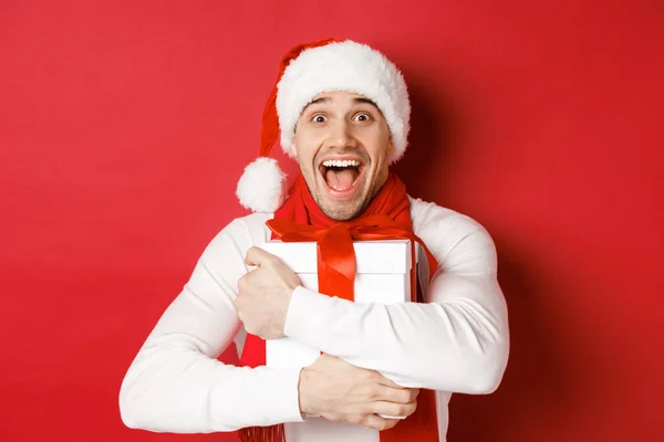 Begreppet vinter semester, jul och livsstil. Närbild av glad man i Santa hatt mottagande närvarande, ser glad och kramas presentförpackning, står över röd bakgrund — Stockfoto