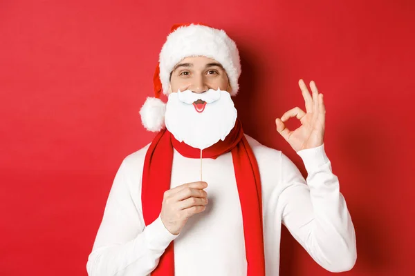 Концепция Рождества, зимних праздников и празднования. Приятно красивый мужчина в шляпе Санты, держа длинную белую бороду маску и показывая хорошо знак, стоя на красном фоне — стоковое фото