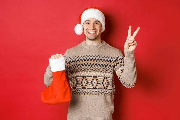 Conceito de férias de inverno, ano novo e celebração. Imagem de homem sorridente feliz em chapéu de santa e suéter, mostrando sinal de paz e uma bolsa de Natal com presentes, fundo vermelho — Fotografia de Stock