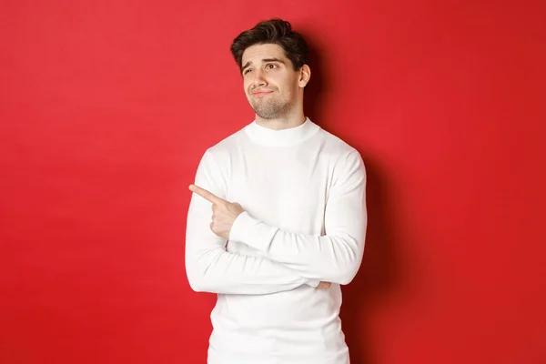 Begrepet vinterferie. Bilde av misfornøyd ung mann i hvit genser, som ser og peker til venstre på noe gjennomsnittlig, står skeptisk til rød bakgrunn – stockfoto
