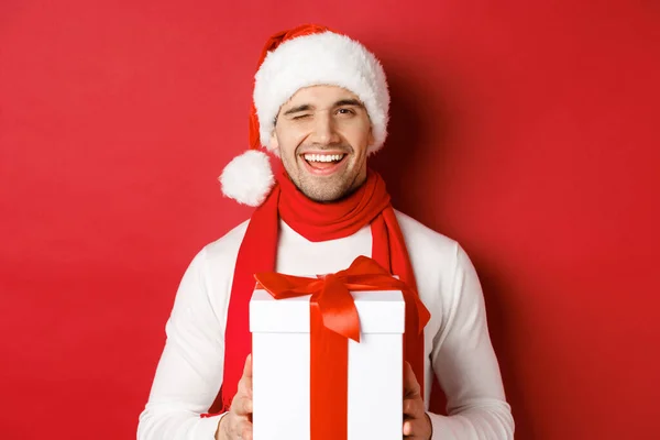 Conceito de férias de inverno, Natal e estilo de vida. Bonito homem atrevido em santa chapéu e cachecol, segurando presente e sorrindo, piscando para a câmera, de pé sobre o fundo vermelho — Fotografia de Stock