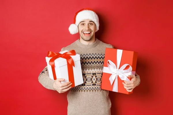 Imagen de chico atractivo alegre sosteniendo regalos de Navidad, de pie en sombrero de santa y suéter de invierno, sonriendo feliz, de pie sobre fondo rojo — Foto de Stock