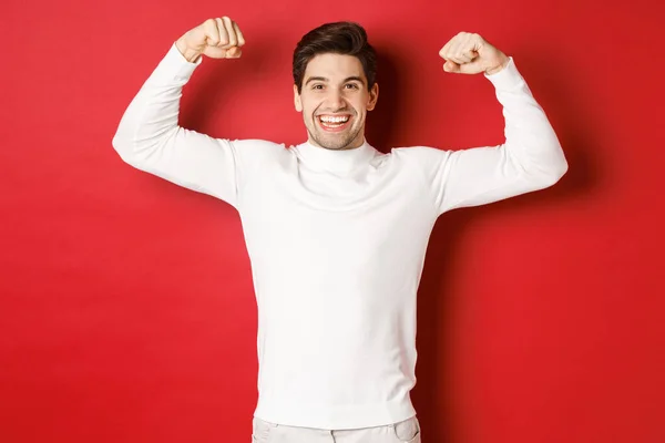 Retrato de hombre guapo sonriente en suéter blanco, bíceps flexionantes y jactancia con fuerza, músculos fuertes después del entrenamiento, de pie sobre fondo rojo —  Fotos de Stock