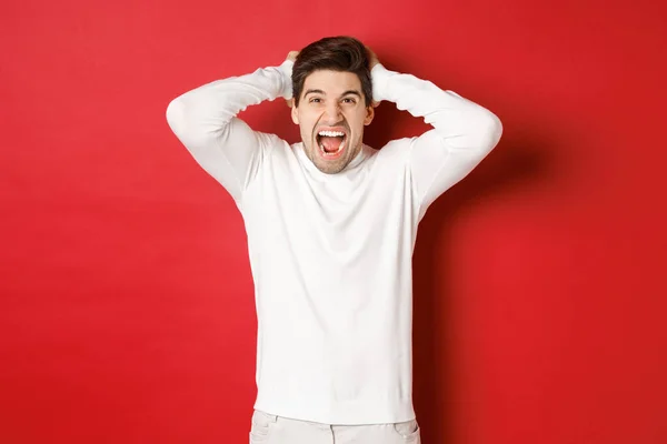 Portrait of frustrated man in white sweater, shouting in anger, holding hands on head and grimacing, feeling distressed, standing over red background — Stock Photo, Image