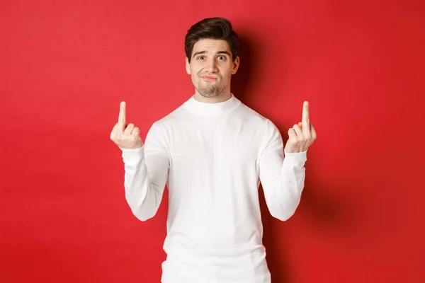 Image of pissed-off and distressed man telling to fuck off, showing middle-fingers and looking upset, standing over red background in white sweater —  Fotos de Stock