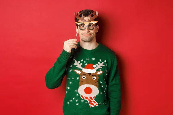Vtipný muž ve vánočním svetru a párty maska, slaví zimní prázdniny, ukazuje legrační tváře, stojí proti červenému pozadí — Stock fotografie