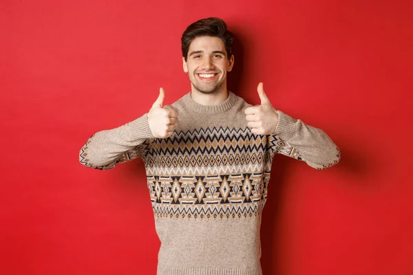 Retrato de chico guapo feliz y complacido en suéter de Navidad, mostrando los pulgares hacia arriba y asiente con la cabeza en la aprobación, sonriendo satisfecho, de pie sobre el fondo rojo —  Fotos de Stock