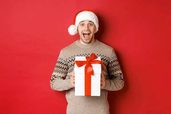 Imagen de hombre guapo emocionado recibiendo regalo de Navidad, usando sombrero de santa y suéter de invierno, gritando de alegría, sosteniendo presente y de pie sobre fondo rojo — Foto de Stock