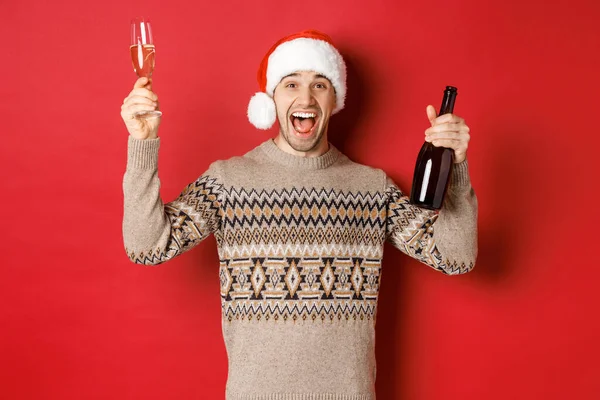 Koncepcja ferii zimowych, świąt i uroczystości. Podniecony przystojniak w swetrze i kapeluszu Mikołaja, cieszący się imprezą noworoczną, podnoszący szklankę i butelkę szampana, bawiący się — Zdjęcie stockowe