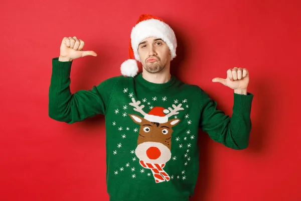 Beeld van knappe en zelfverzekerde jongeman in groene trui en kerstmuts, wijzend naar zichzelf, kerstviering, staande over rode achtergrond — Stockfoto