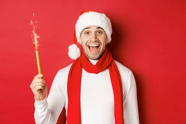 Konzept von Weihnachten, Winterurlaub und Feiern. Schöner Mann feiert Neujahr und hat Spaß, hält Wunderkerze in der Hand und lächelt, trägt Weihnachtsmütze, steht über rotem Hintergrund — Stockfoto