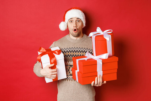 Concept of winter holidays, new year and celebration. Image of surprised handsome guy in sweater and santa hat, receiving gifts, holding boxes with presents and looking amazed