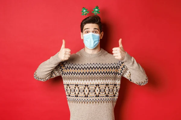 Conceito de ano novo, covid-19 e distanciamento social. Homem caucasiano alegre em suéter e máscara médica, celebrando o Natal durante a pandemia, mostrando polegar para cima, fundo vermelho — Fotografia de Stock