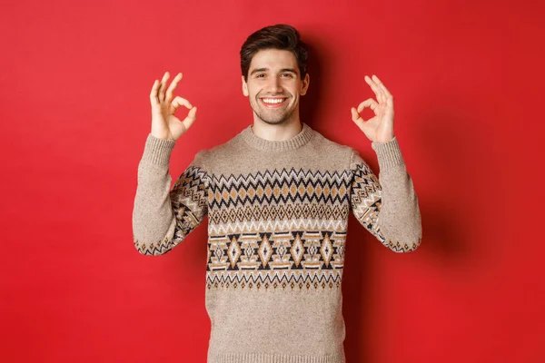Begrepet julefeiring, vinterferier og livsstil. Tilfreds kjekk mann i genser, viser ok tegn og smiler fornøyd, anbefaler noe, rød bakgrunn – stockfoto