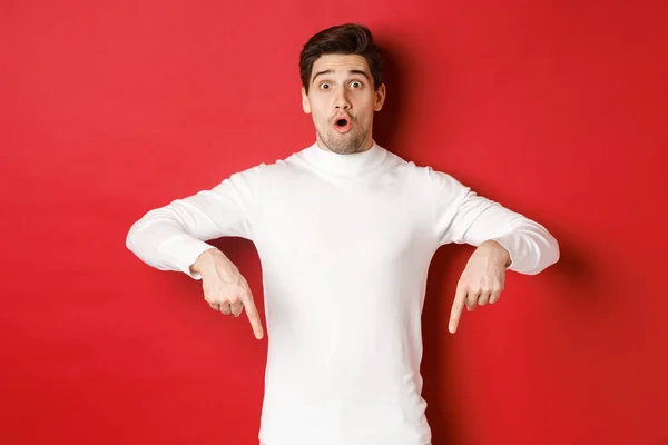 Imagem de cara atraente surpreso em suéter branco, mostrando anúncio, apontando os dedos para baixo e olhando espantado, de pé sobre fundo vermelho — Fotografia de Stock