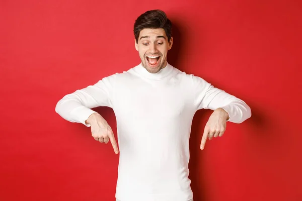 Portrait of happy good-looking man, pointing fingers down and looking at advertisement with excited smile, standing over red background — Stock Photo, Image