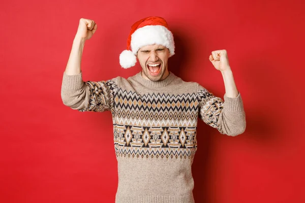 Hombre guapo satisfecho, sintiéndose afortunado y feliz, gritando de alegría y haciendo bombas de puño, celebrando la victoria o ganar, recibir regalo de Navidad impresionante, de pie en sombrero de santa sobre fondo rojo — Foto de Stock
