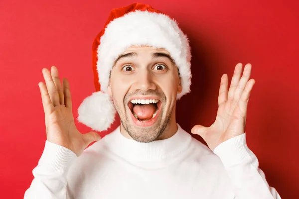 Närbild av glad ung man i Santa hatt, gör stora jul tillkännagivande, leende förvånad, står över röd bakgrund — Stockfoto