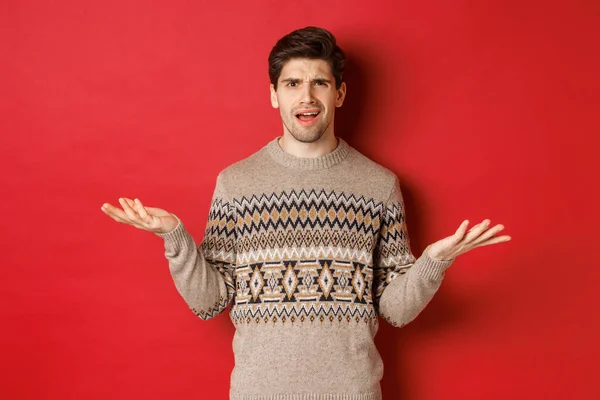 Retrato de chico guapo confundido y decepcionado, quejándose de la Navidad, las manos extendidas hacia los lados y frunciendo el ceño disgustado, de pie en suéter de Navidad sobre fondo rojo —  Fotos de Stock