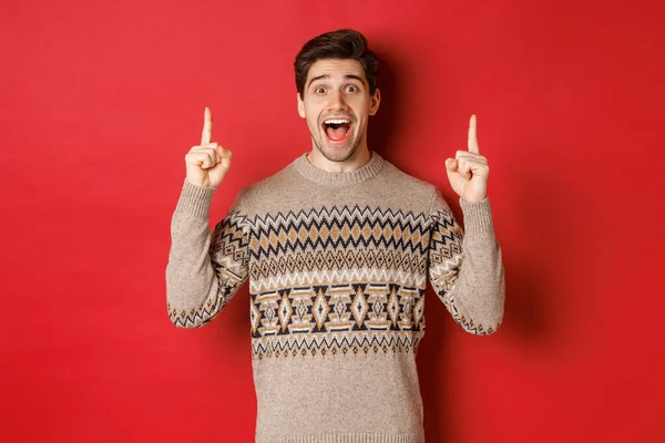 Изображение красивого взволнованного мужчины, счастливого от рождественских праздников, указывающего пальцем вверх и выглядящего веселым, показывающего рекламу, стоящего в свитере на красном фоне — стоковое фото