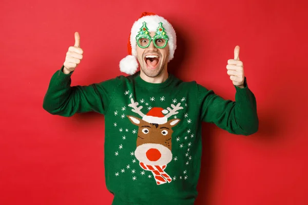 Portret super szczęśliwego młodzieńca w okularach imprezowych, czapce Mikołaja i swetrze, z kciukami do góry w aprobacie, polecający noworoczną ofertę promocyjną, stojący nad czerwonym tłem — Zdjęcie stockowe