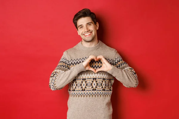 Képzeld el a jóképű szakállas fickót téli pulóverben, boldog karácsonyt kívánva és szívjelet mutatva, kedves mosollyal a kamera előtt, vörös háttér előtt — Stock Fotó