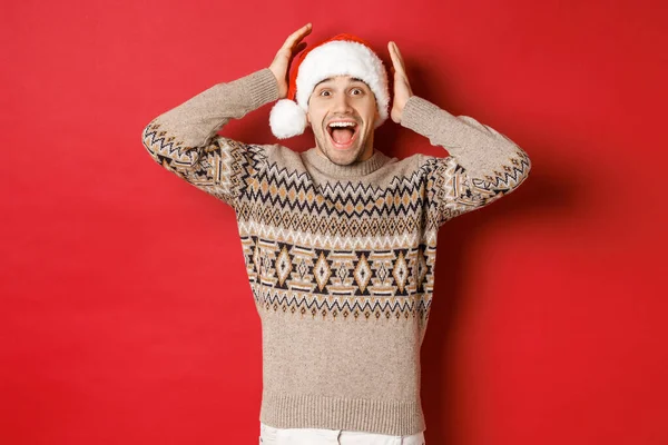 Bild eines überraschten und glücklichen jungen Mannes mit Weihnachtsmütze und Weihnachtspullover, der ein tolles Geschenk erhält und aufgeregt vor rotem Hintergrund steht — Stockfoto