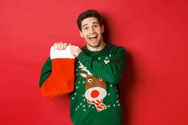 Obrázek atraktivního veselého muže ve svetru, držící vánoční punčochu s dárky, oslavující zimní prázdniny, stojící nad červeným pozadím — Stock fotografie