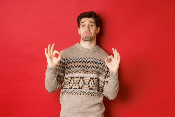 Hombre guapo impresionado en suéter de Navidad, mostrando signos de bien y alabando buena fiesta, asintiendo con la cabeza en la aprobación, de pie complacido contra el fondo rojo —  Fotos de Stock
