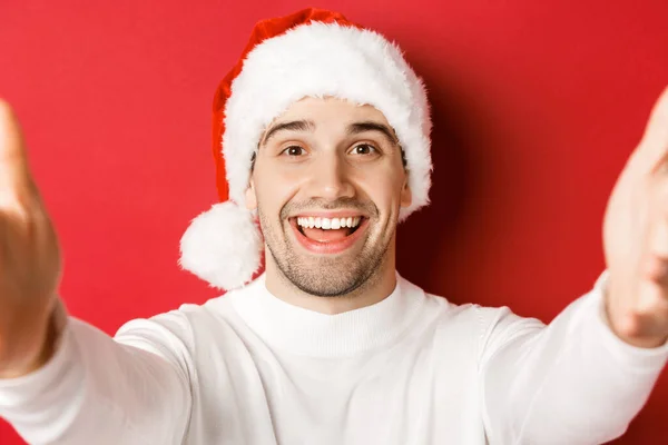 Close-up de homem bonito em Papai Noel, tomando selfie ou ter chamada de vídeo, segurando câmera com ambas as mãos, de pé sobre fundo vermelho — Fotografia de Stock