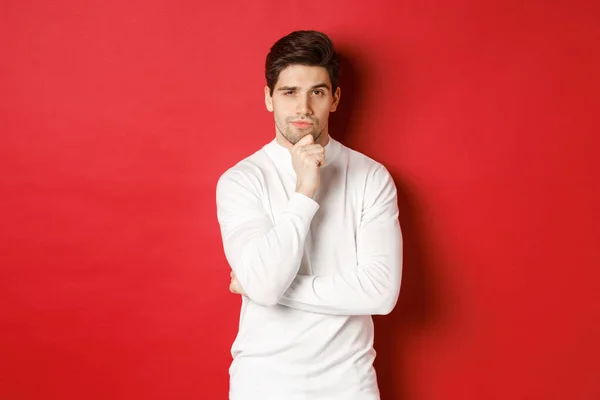 Bilde av omtenksom, kjekk mann som gjør antagelser, tenker og ser på kamera, står i hvit genser mot rød bakgrunn – stockfoto
