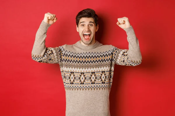 Imagen de chico guapo feliz regocijándose en la víspera de Navidad, levantando las manos y gritando de alegría sí, lograr la meta de año nuevo, de pie sobre el fondo rojo en suéter —  Fotos de Stock