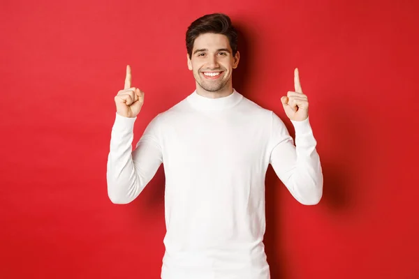 Koncepcja ferii zimowych. Przystojny młody człowiek z włosia, ubrany w biały sweter, pokazujący świąteczną reklamę na przestrzeni kopiowania, wskazujący palcem w górę i uśmiechniętym, czerwonym tle — Zdjęcie stockowe