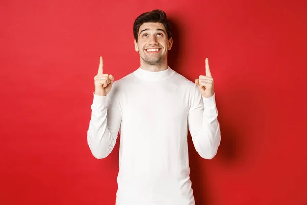 Conceito de férias de inverno. Imagem de homem atraente feliz em camisola branca, sorrindo enquanto apontando e olhando para o espaço de cópia no fundo vermelho — Fotografia de Stock