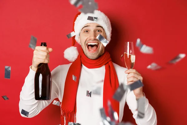 Begreppet vinter semester, jul och livsstil. Närbild av glad man firar nytt år, håller champagne flaska och glas, står över röd bakgrund med konfetti — Stockfoto