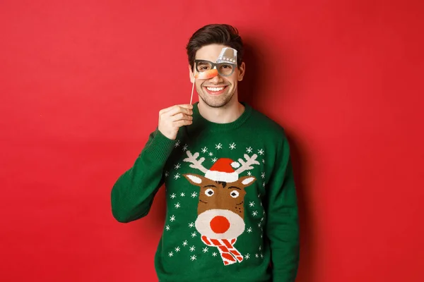 Vtipný muž v vánoční svetr a party maska, slaví zimní prázdniny, šťastný úsměv, stojící nad červeným pozadím — Stock fotografie