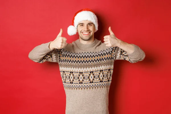 Портрет привабливого щасливого чоловіка в капелюсі Санти і светрі, показує великі пальці в схваленні і посмішці, бажаючи веселого Різдва, стоячи на червоному тлі — стокове фото