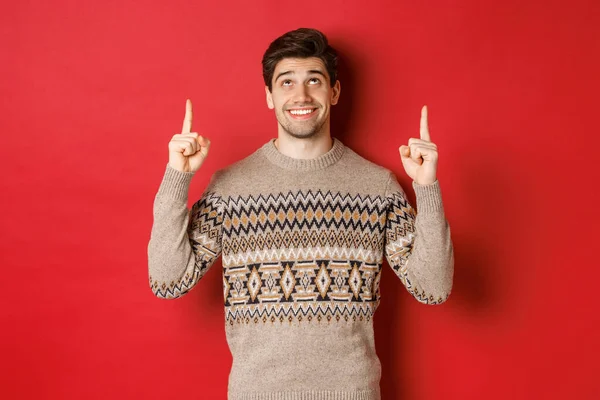 Портрет мечтательного красавца, который думает о рождественских праздниках, носит зимний свитер, показывает и смотрит вверх на новогоднее промо-предложение, стоя на красном фоне — стоковое фото