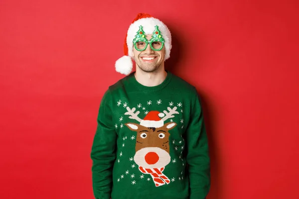 Yeşil Noel süveteri, Noel Baba şapkası ve parti gözlüğü giymiş yakışıklı bir adamın portresi. Mutlu gülümsüyor, kırmızı arka planda duruyor. — Stok fotoğraf