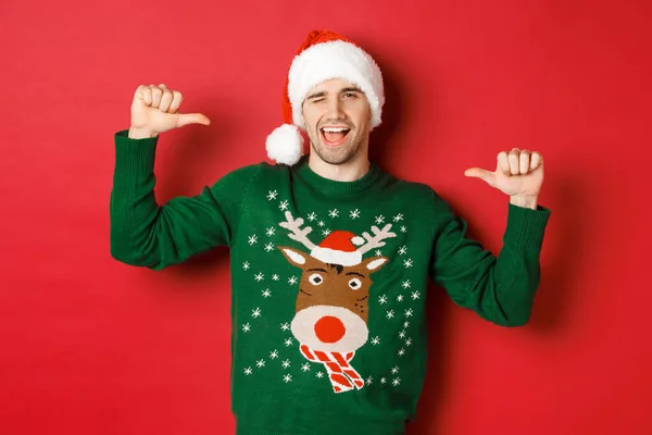 Concepto de vacaciones de invierno, Navidad y estilo de vida. Hombre guapo descarado en sombrero de santa y suéter verde, apuntándose a sí mismo y guiñando el ojo, de pie sobre el fondo rojo —  Fotos de Stock