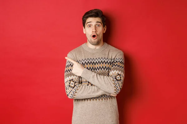 Imagem de cara bonito impressionado, mostrando incrível oferta promo natal, apontando o dedo no canto superior esquerdo e olhando espantado, de pé em xmas suéter sobre fundo vermelho — Fotografia de Stock