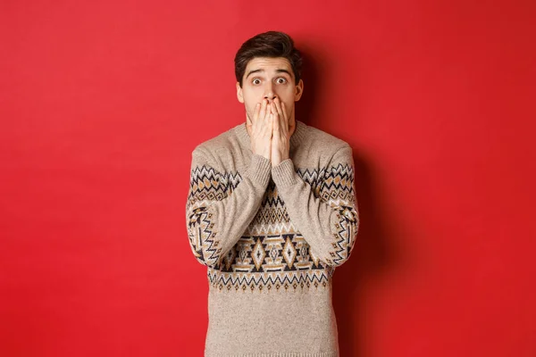 Портрет шокированного и безмолвного белого парня в рождественском свитере, реагирующего на объявление, стоящего испуганного на красном фоне — стоковое фото